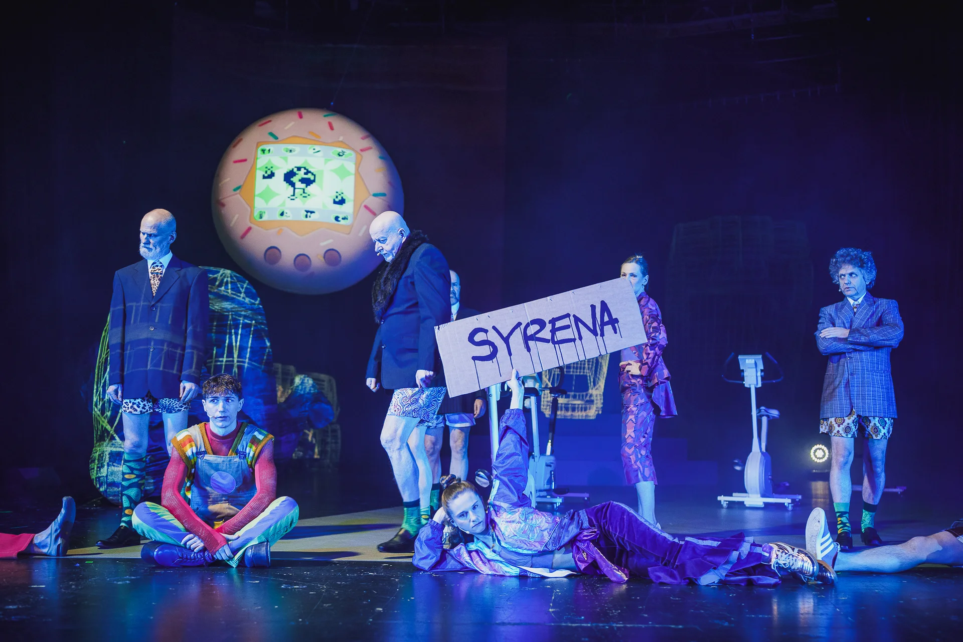 fot. Piotr Nykowski Grupa aktorów na scenie. Na pierwszym planie leży kobieta trzymająca transparent z napisem "syrena". Po jej prawej stronie siedzi mężczyzna ze skrzyżowanymi nogami. Na drugim planie znajdują się mężczyźni w marynarkach i krótkich spodenkach oraz kobieta we wzorzystym garniturze. W tle widać podwieszoną wielką kulę, na której wyświetlane jest różowe tamagotchi.