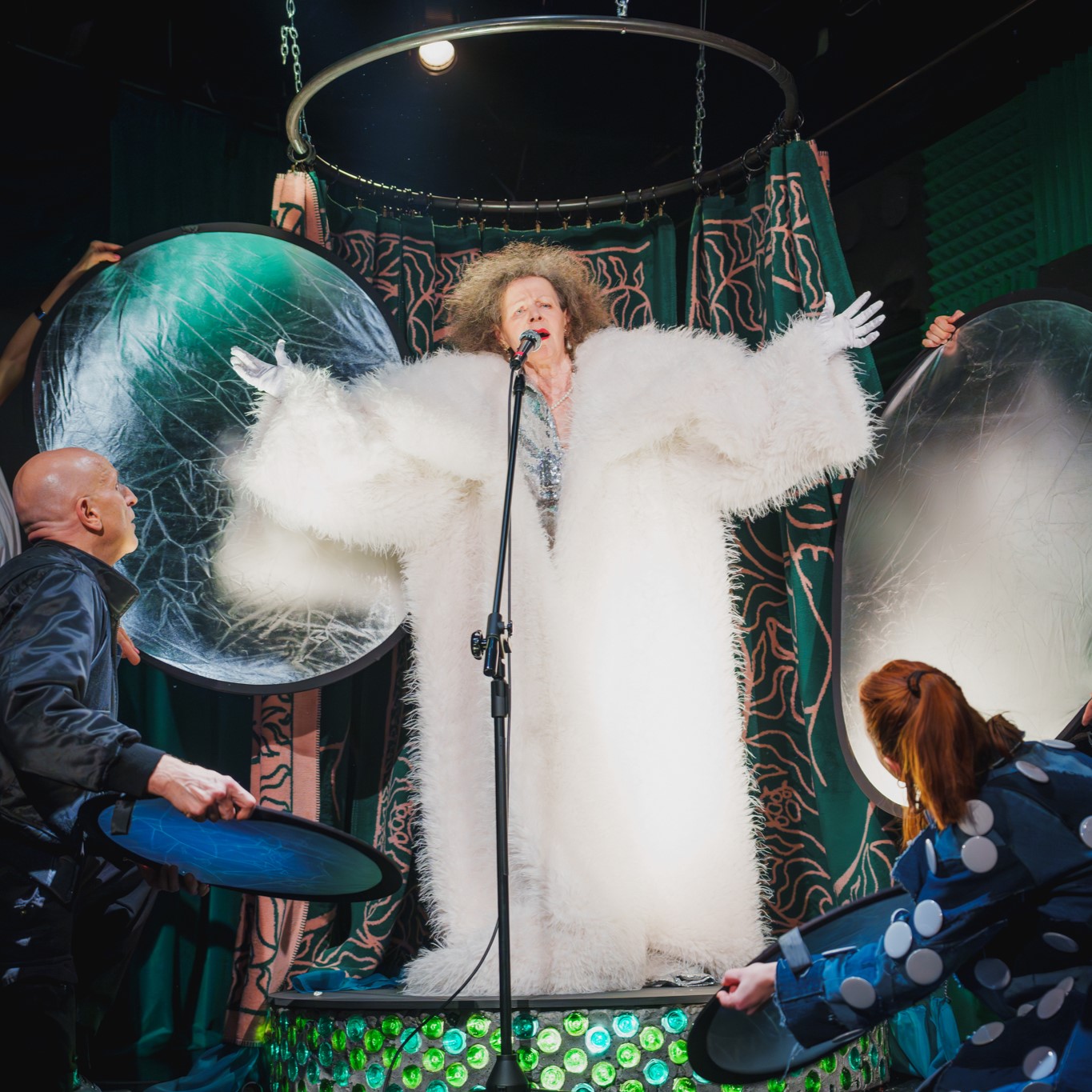 Aktorka ubrana w duże białe futro i srebrne rękawiczki. Stoi przy mikrofonie. Śpiewa. Dookoła niej grupa aktorów i aktorek trzyma panele odbijające światło.