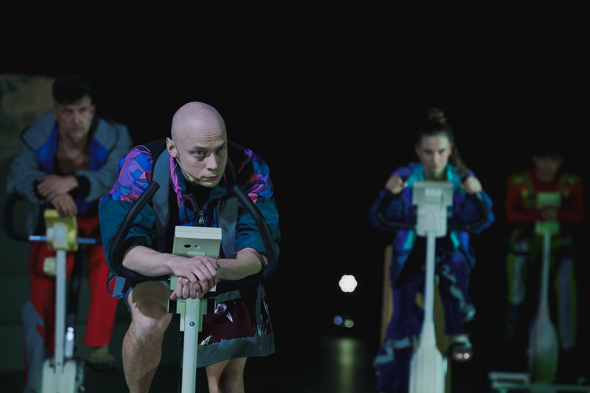 „Sztuka protestu”, fot. Piotr Nykowski Aktor ubrany w kolorowy dres jedzie na rowerze treningowym. W tle widać dwóch aktorów i aktorkę również na rowerach.