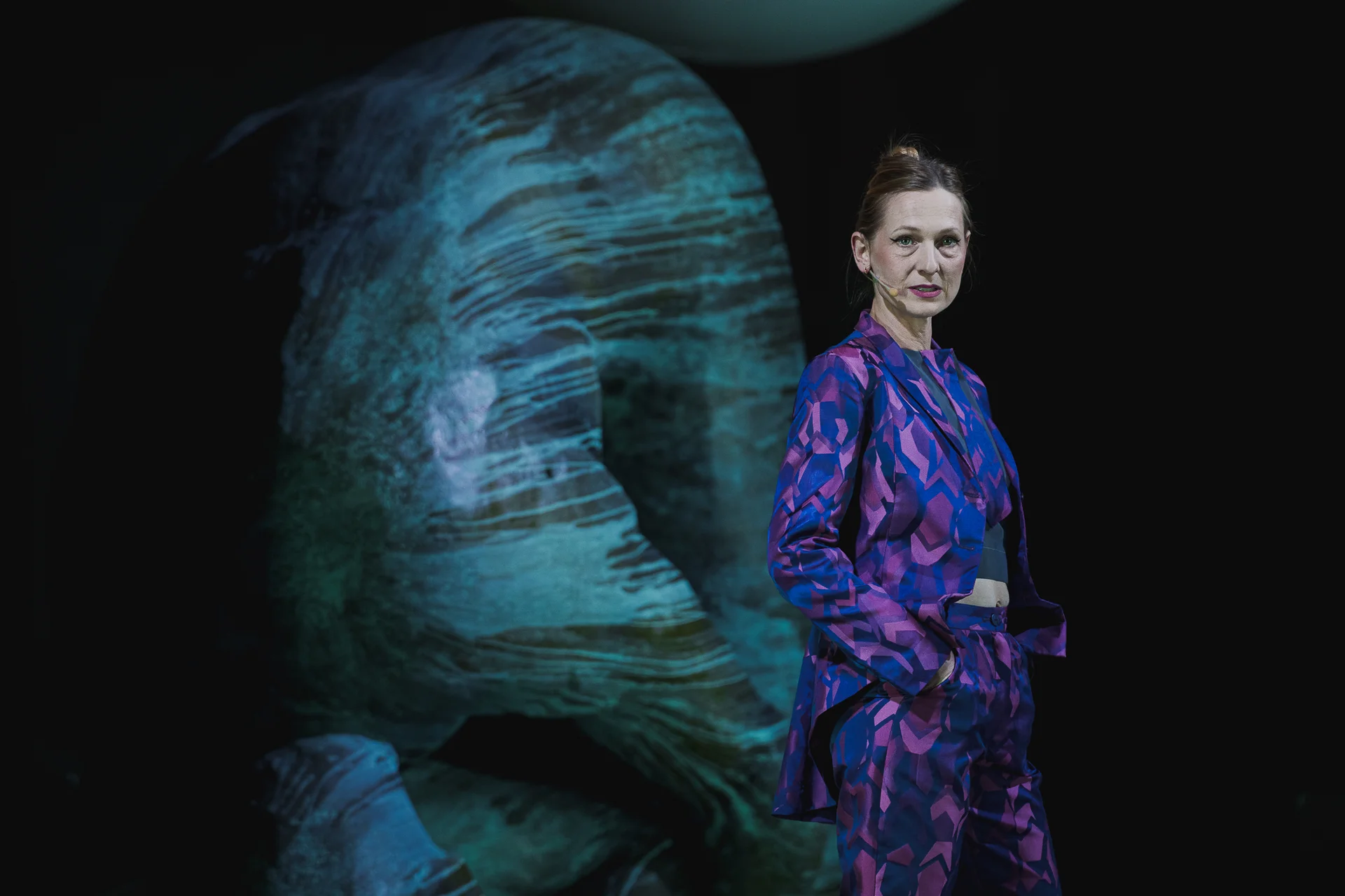 Aktorka ubrana w kolorowy garnitur stoi na tle dużej rzeźby. 