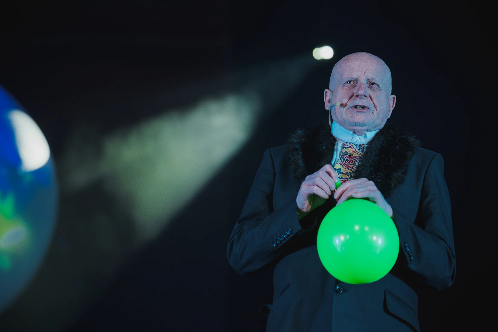 Aktor ubrany w czarną marynarkę z futrzanym kołnierzem. W dłoniach trzyma zielony balonik. 