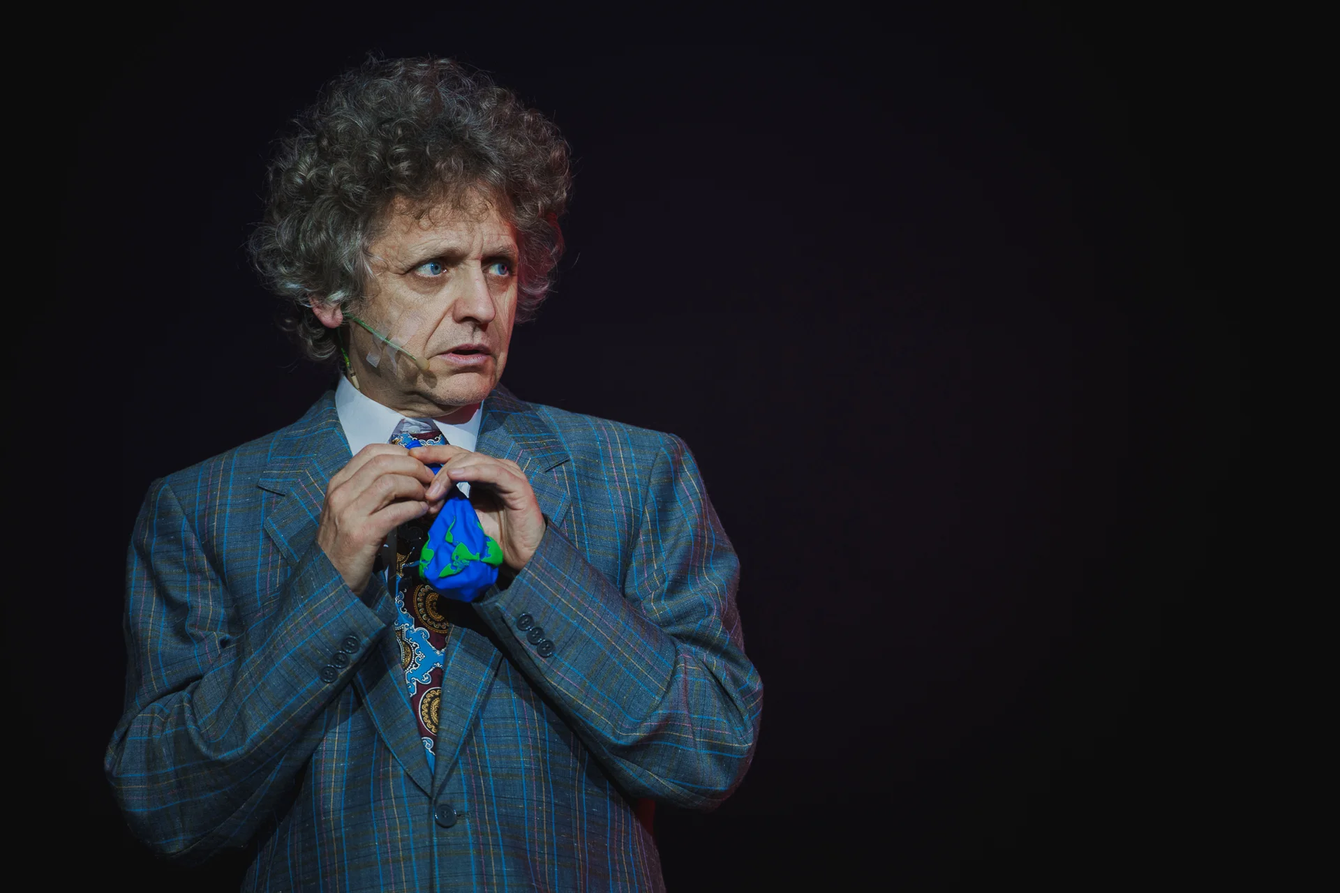 Aktor ubrany w szarą marynarkę. W dłoniach trzyma mały niebieski balonik. Z przestraszoną miną patrzy w lewo. 