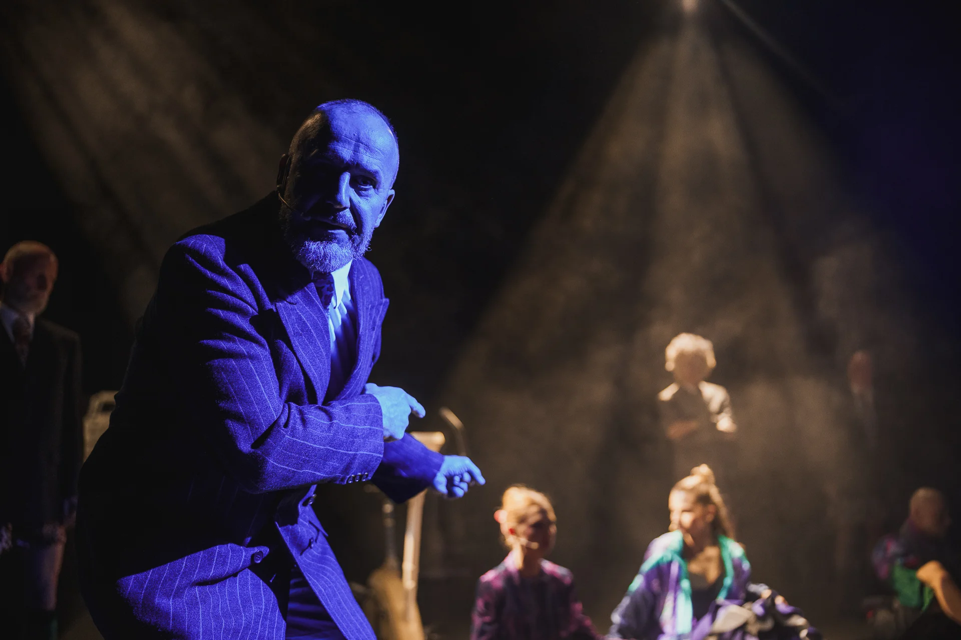 Aktor ukazany w niebieskim świetle. Ubrany w marynarkę. Wskazuje na aktorów w tle. 