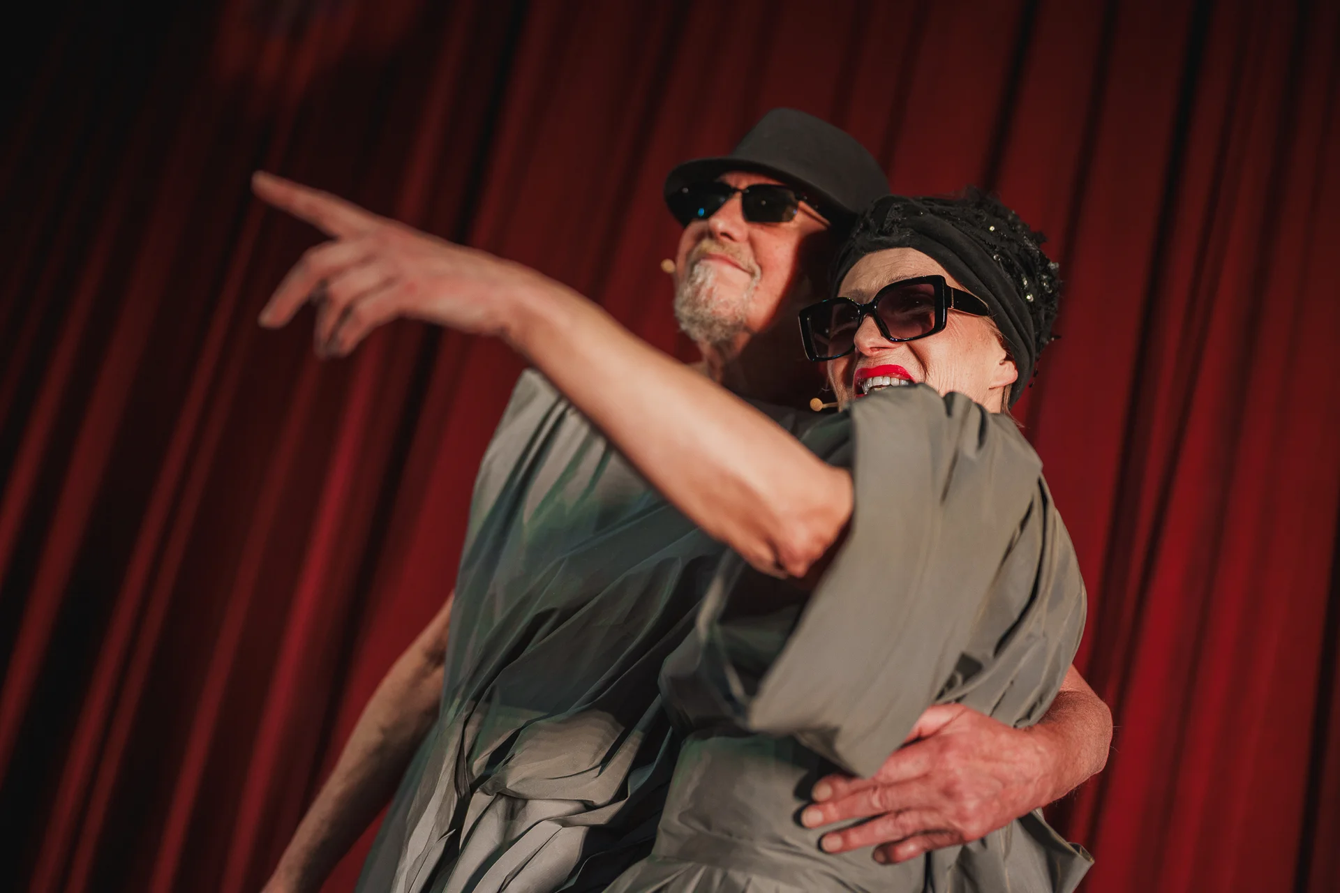 Aktorka i aktor ubrani w szare drapowane szaty. Na głowach mają ciemne kapelusze i przeciwsłoneczne okulary. Obejmują się. Kobieta wskazuje przed siebie prawą ręką. W tle czerwona kurtyna. 