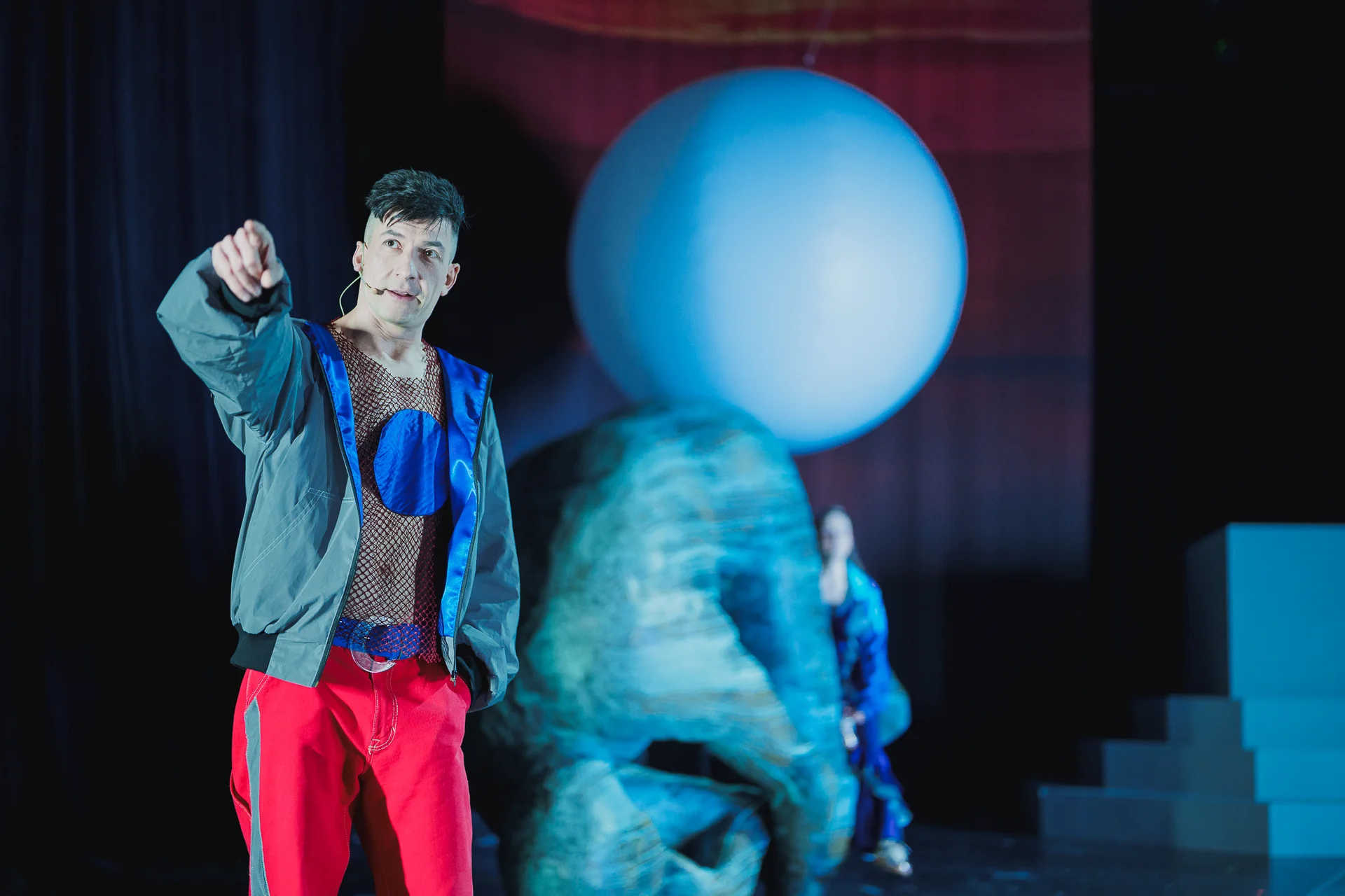 Aktor ubrany w szarą kurtkę sportową i czerwone spodnie. Prawą ręką wskazuje przed siebie. W tle widać dużą rzeźbę.