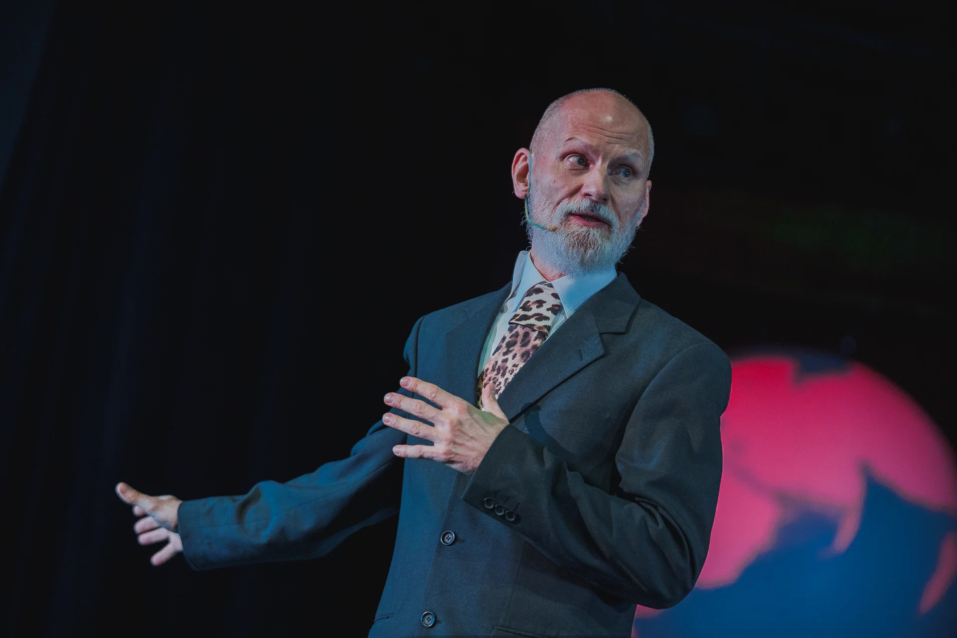 Aktor ubrany w szarą marynarkę. Gestykuluje wskazując w prawą stronę. 