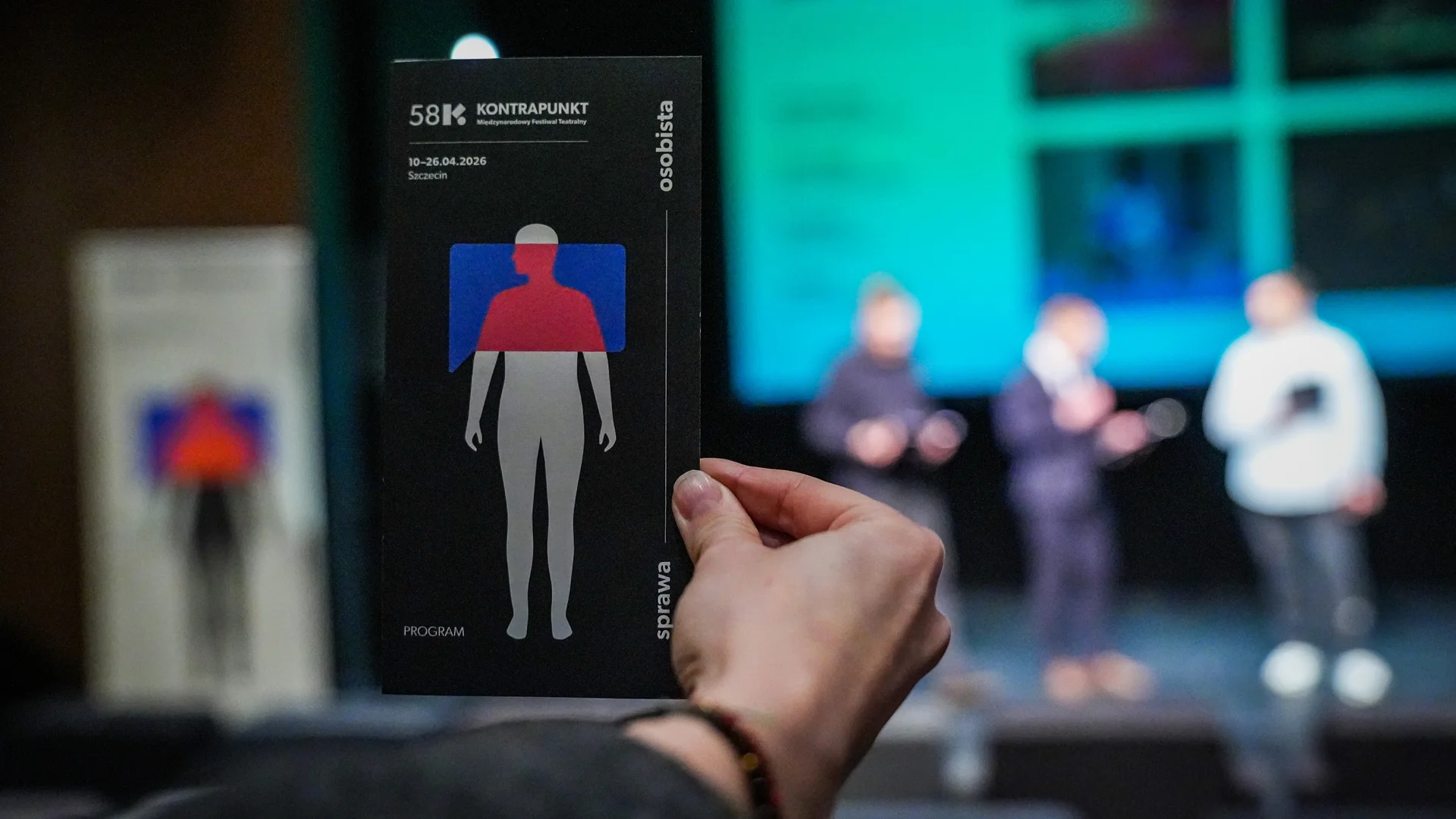 fot. Tomasz Jaworski, Szczecińska Agencja Artystyczna Wyciągnięta przed siebie dłoń, a w niej program festiwalu Kontrapunkt. W tle trzech mężczyzn stojących na tle prezentacji.