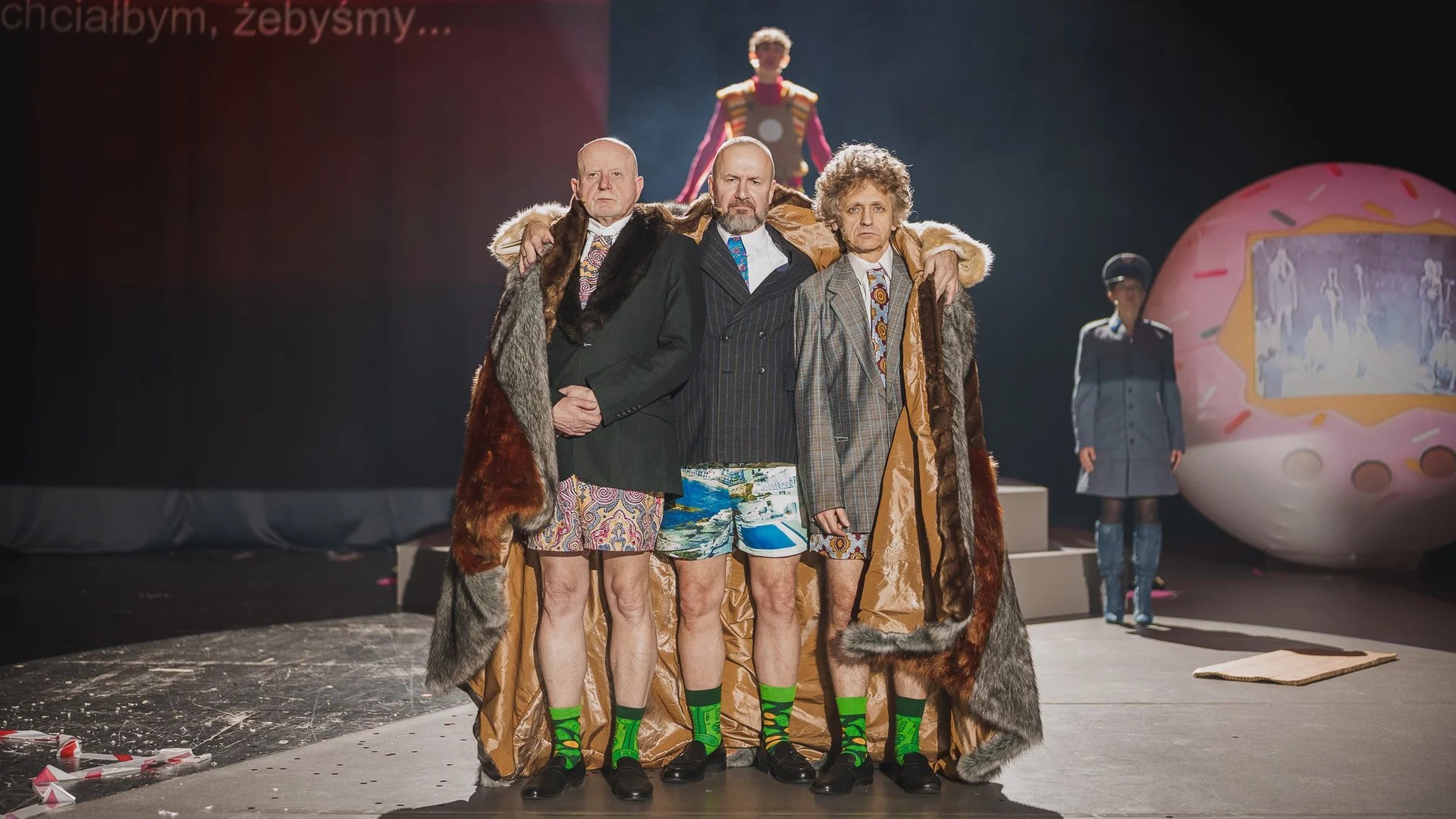 Zdjęcie ze spektaklu Sztuka protestu. Trzech mężczyzn w marynarkach stoi bez spodni. Mają poważne miny, a na sobie krótkie kolorowe spodenki, kolorowe skarpetki. Obejmuje ich trzech wielkie futro.