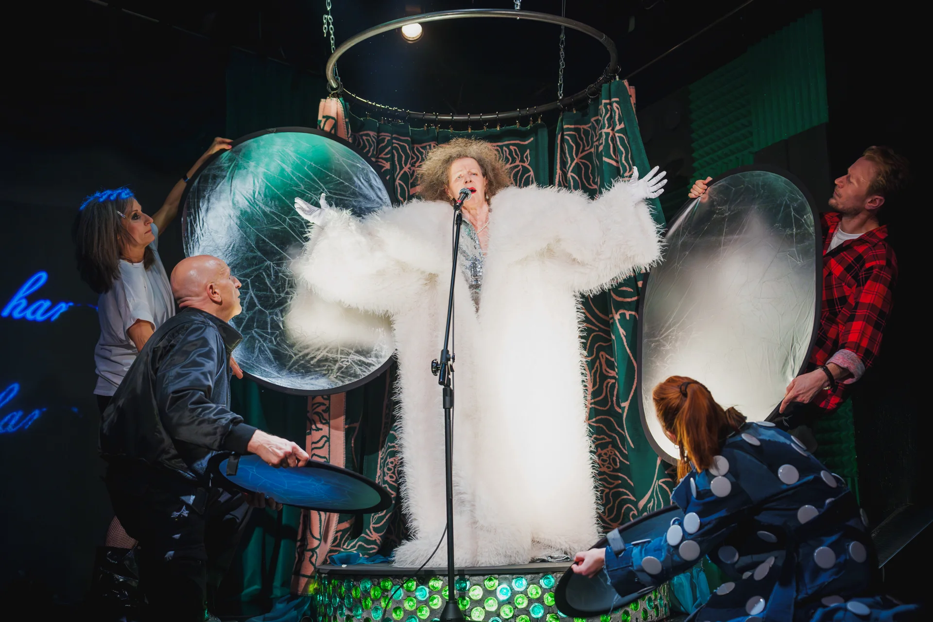 „Piękny i krótki spektakl o komunikacji”, fot. Piotr Nykowski Aktorka ubrana w duże białe futro i srebrne rękawiczki. Stoi przy mikrofonie. Śpiewa. Dookoła niej grupa aktorów i aktorek trzyma panele odbijające światło.