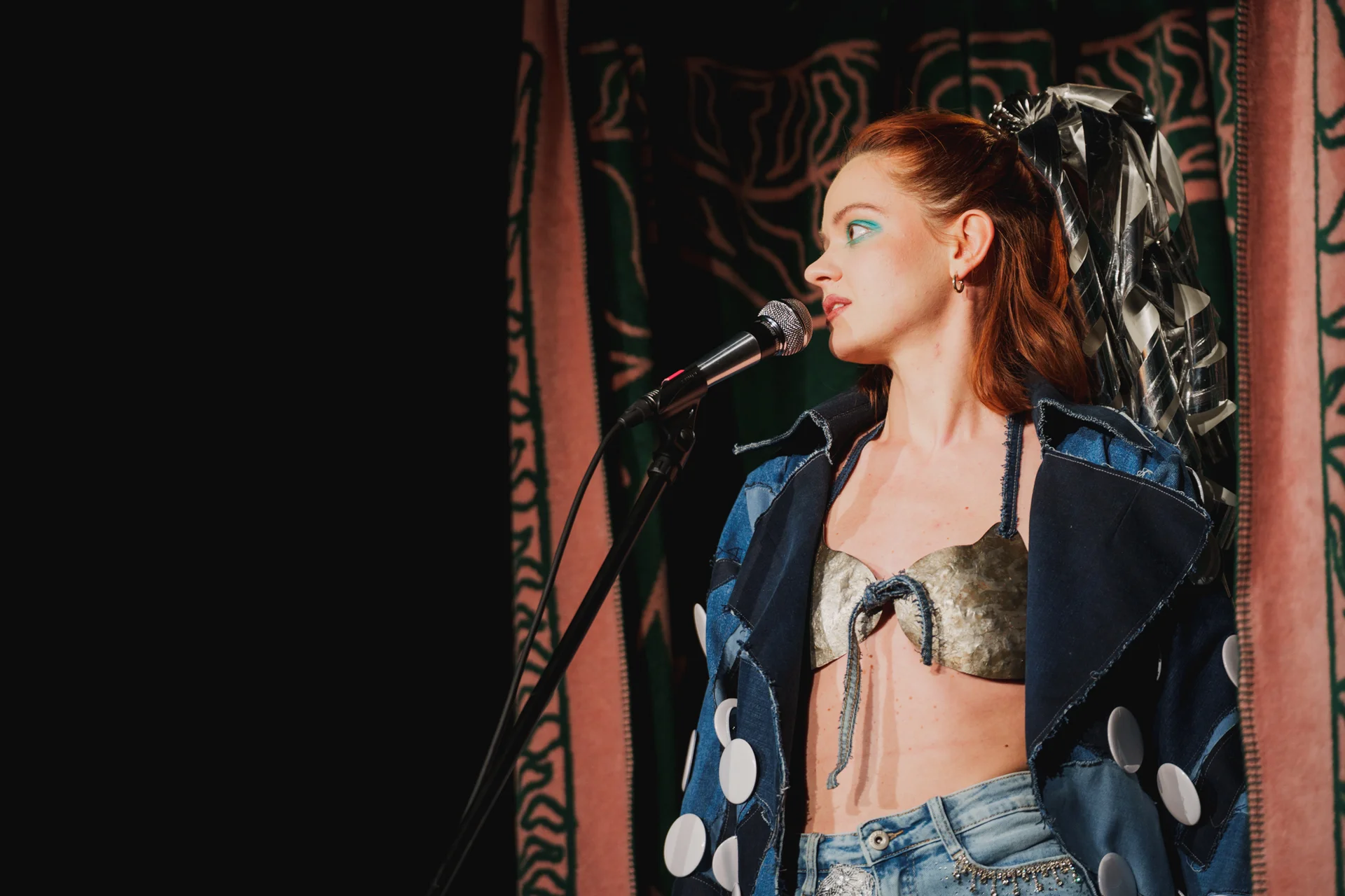 Aktorka w dżinsowym płaszczu i metalowym staniku. Przed nią stoi mikrofon. Spogląda w prawą stronę.