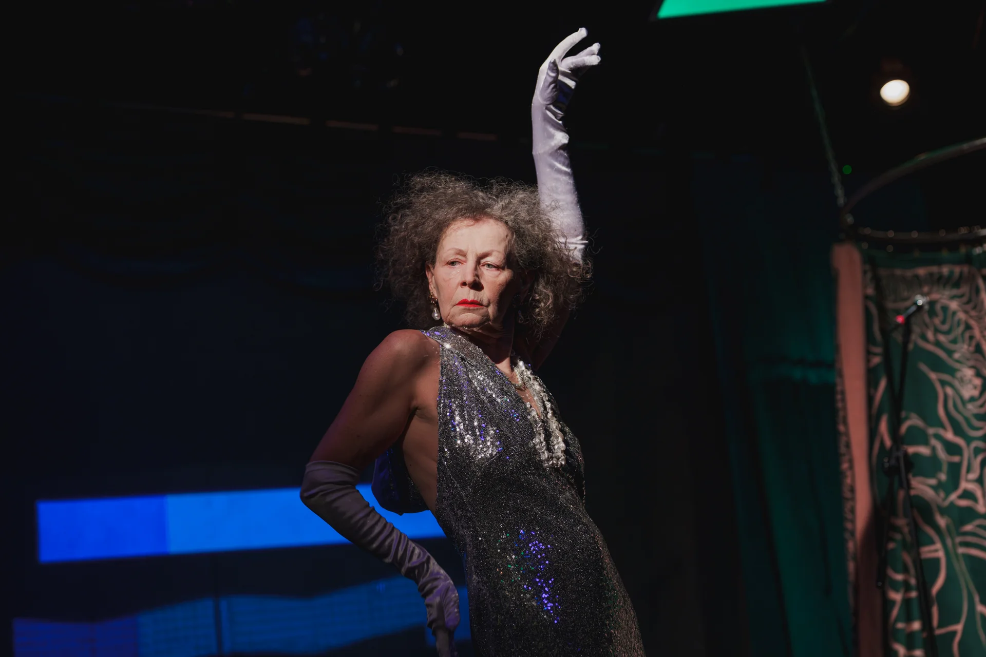 Aktorka pozująca z lewą ręką w górze. Usta ma umalowane czerwoną szminką. Ubrana jest w srebrną suknię.