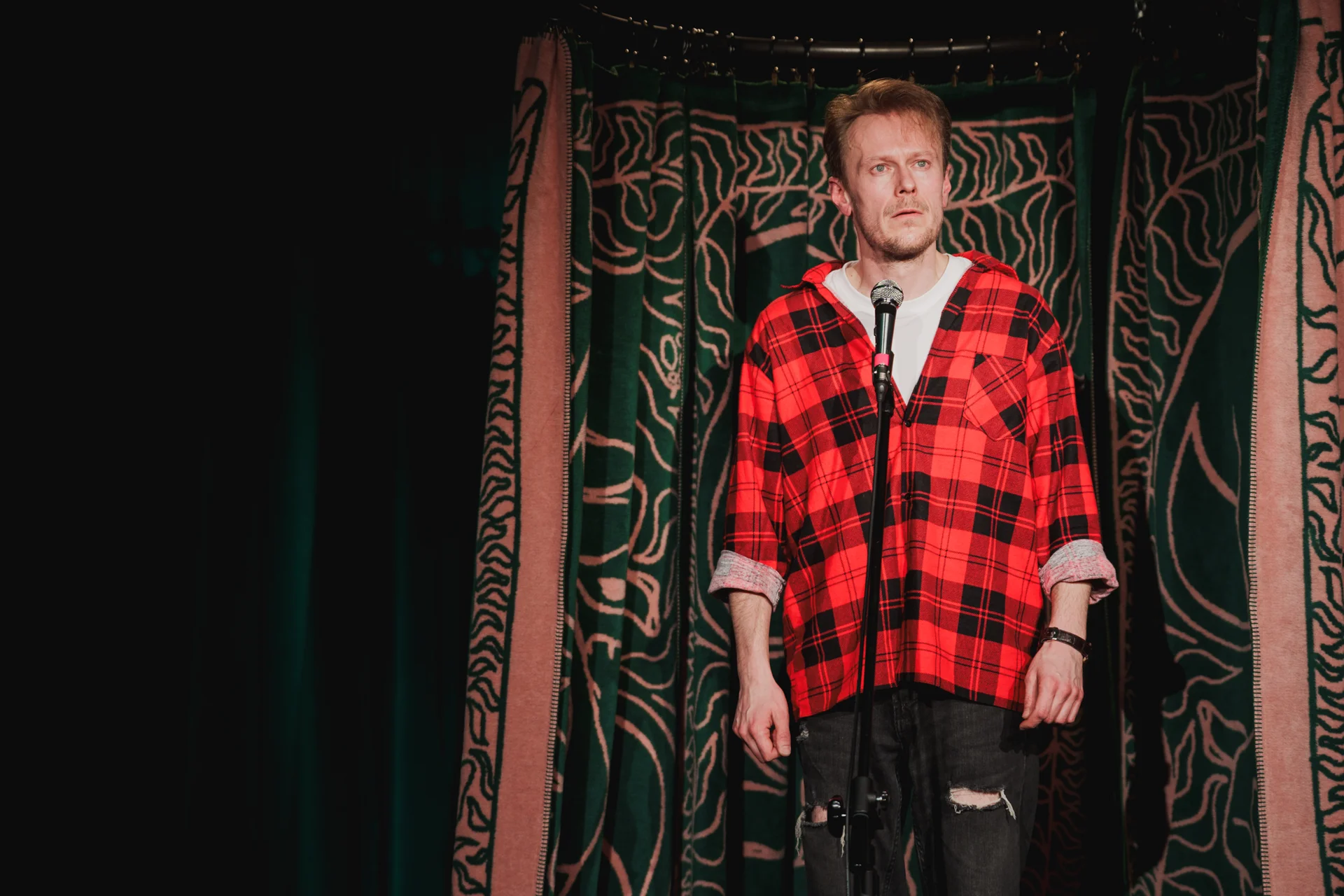 Mężczyzna w czerwonej kraciastej koszuli stoi ze zmartwioną miną za mikrofonem.