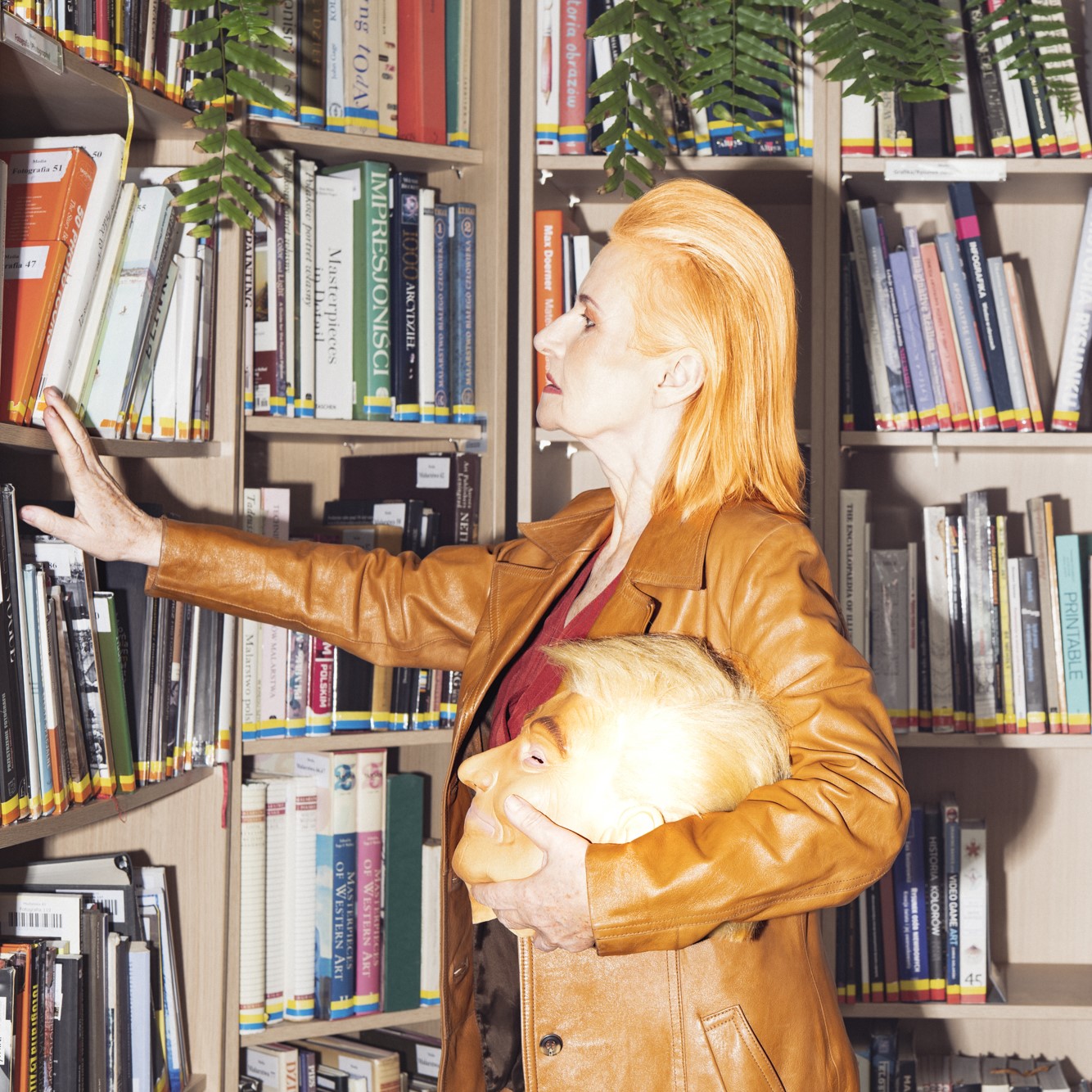 Kobieta o rudych włosach ubrana w brązową skórzaną kurtkę przegląda półki z książkami w bibliotece. W lewej ręce trzyma maskę przedstawiającą Donalda Trumpa.