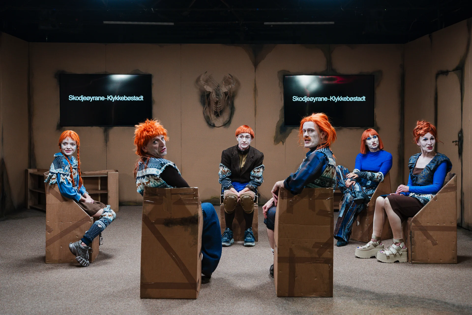 Na kartonowych siedziskach siedzą aktorzy w rudych perukach. Mają na sobie niebieskie kostiumy. Patrzą na publiczność.