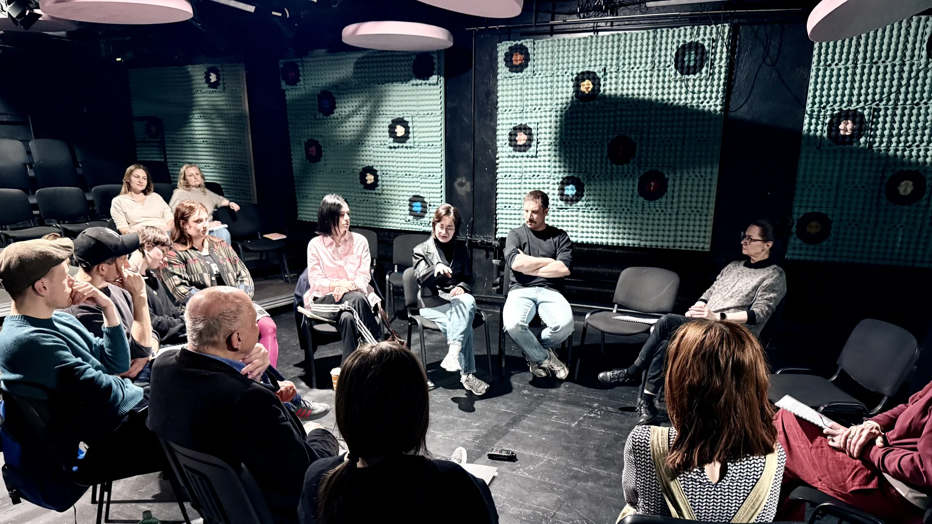 Zdjęcie z pierwszej próby MOZAIKI Zdjęcie z pierwszej próby MOZAIKI. Grupa osób siedząca na krzesłach w kole. Na ścianie zielone kwadraty, z sufitu podwieszane różowe koła.
