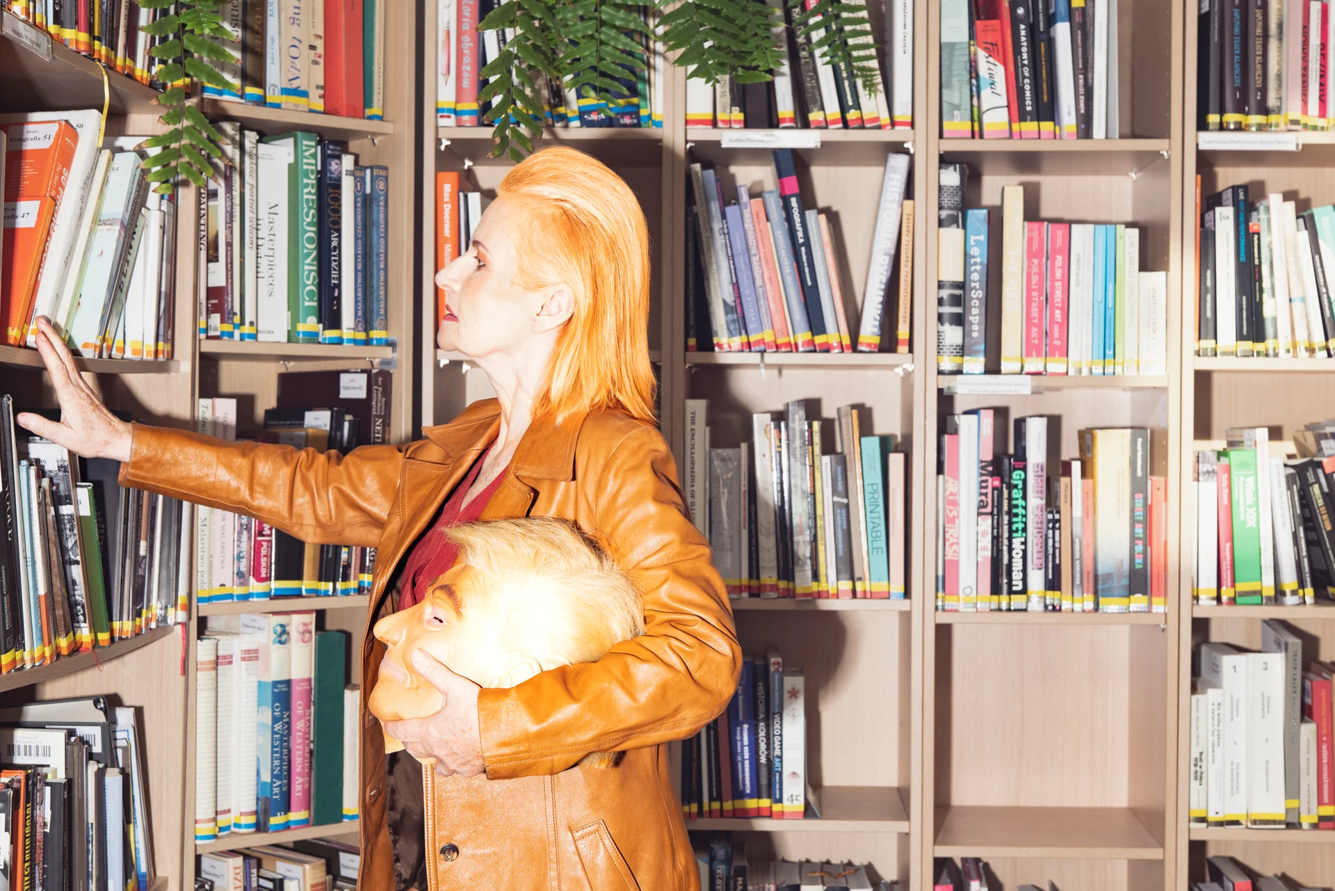 Kobieta o rudych włosach ubrana w brązową skórzaną kurtkę przegląda półki z książkami w bibliotece. W lewej ręce trzyma maskę przedstawiającą Donalda Trumpa.
