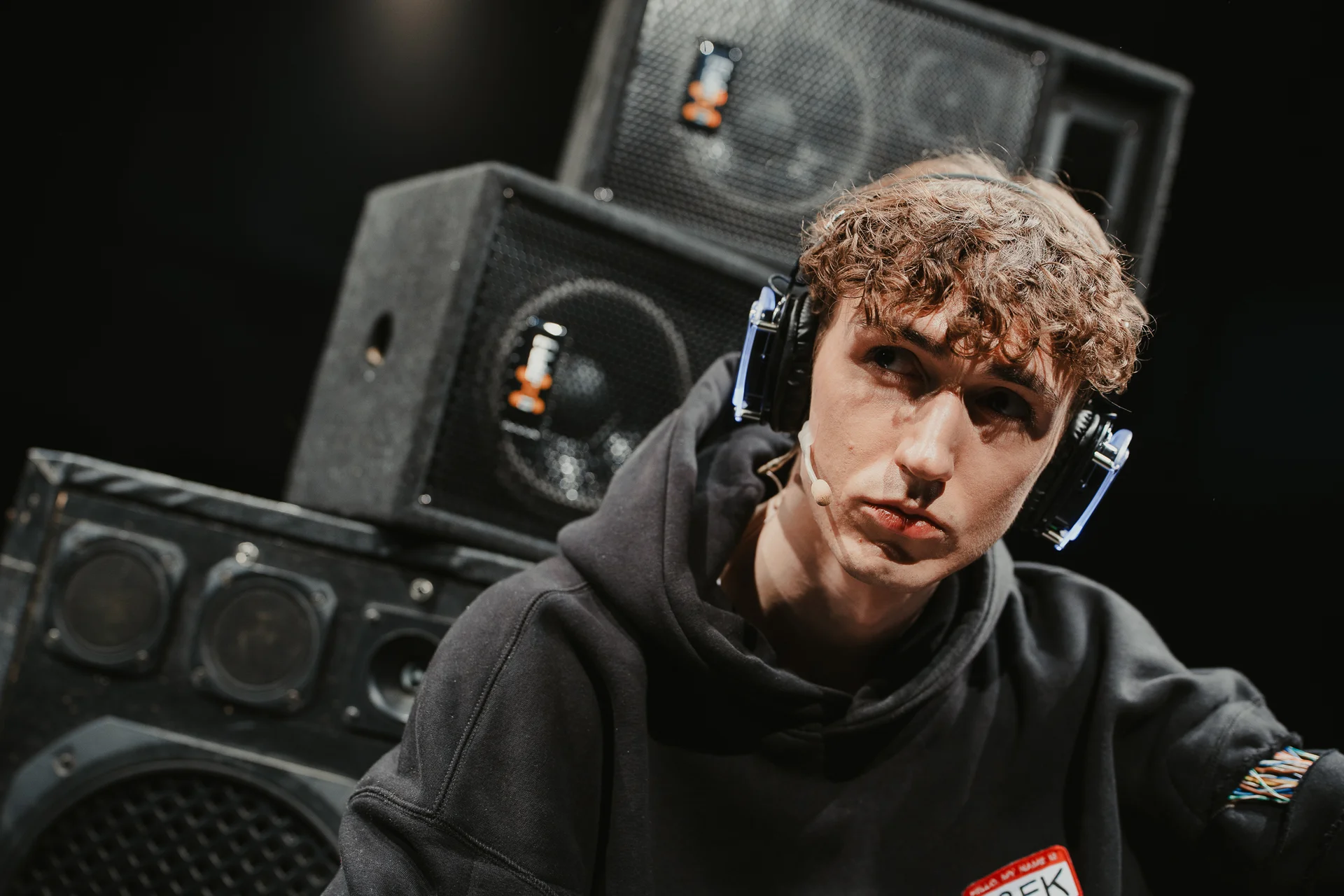 Młody mężczyzna w dużych nausznych słuchawkach, ubrany w czarną bluzę. W tle głośniki.