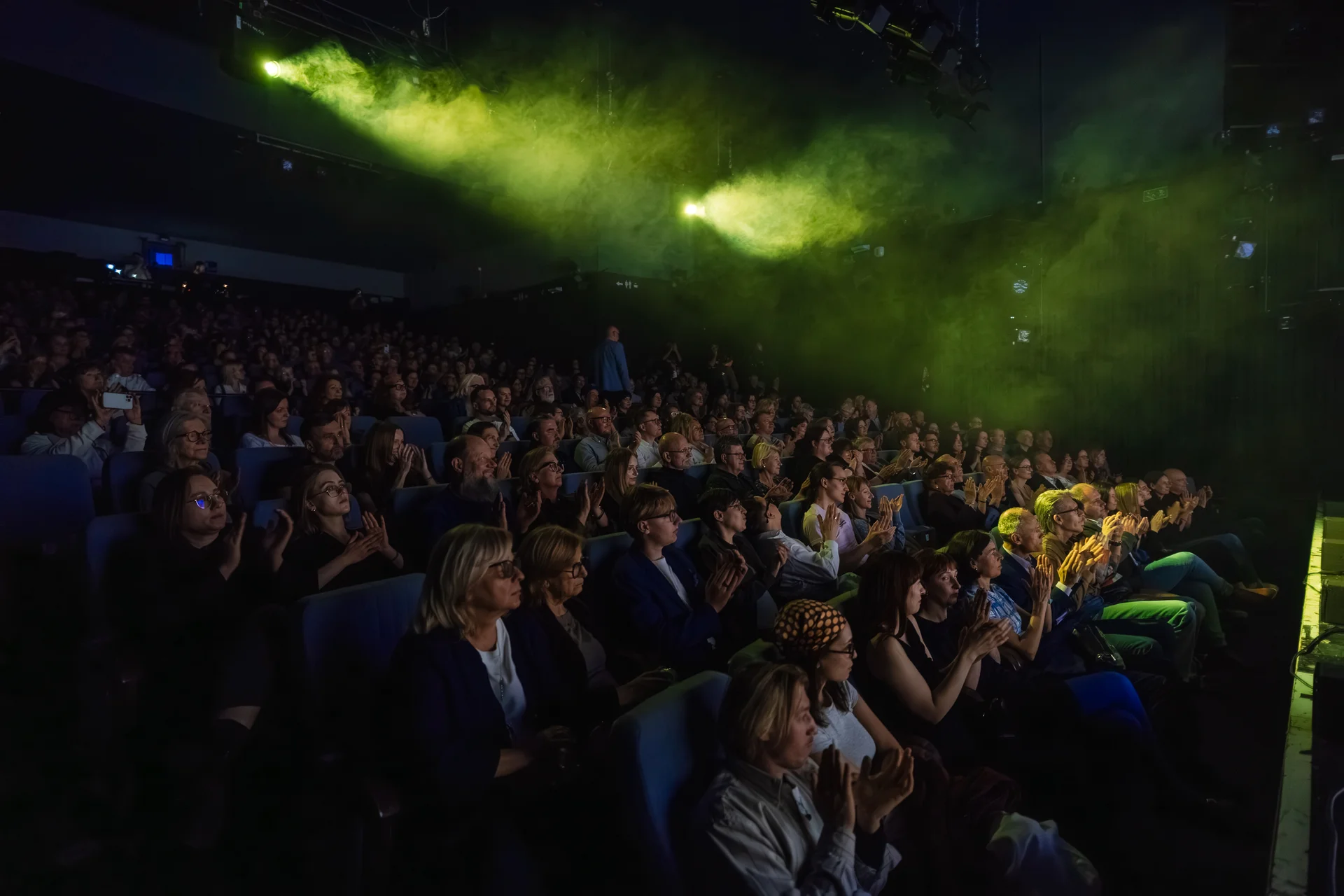 Zdjęcie publiczności festiwalu. Widownia z zainteresowaniem przygląda się scenie, której nie widzimy. Nad nimi unosi się dym oświetlony na zielono.