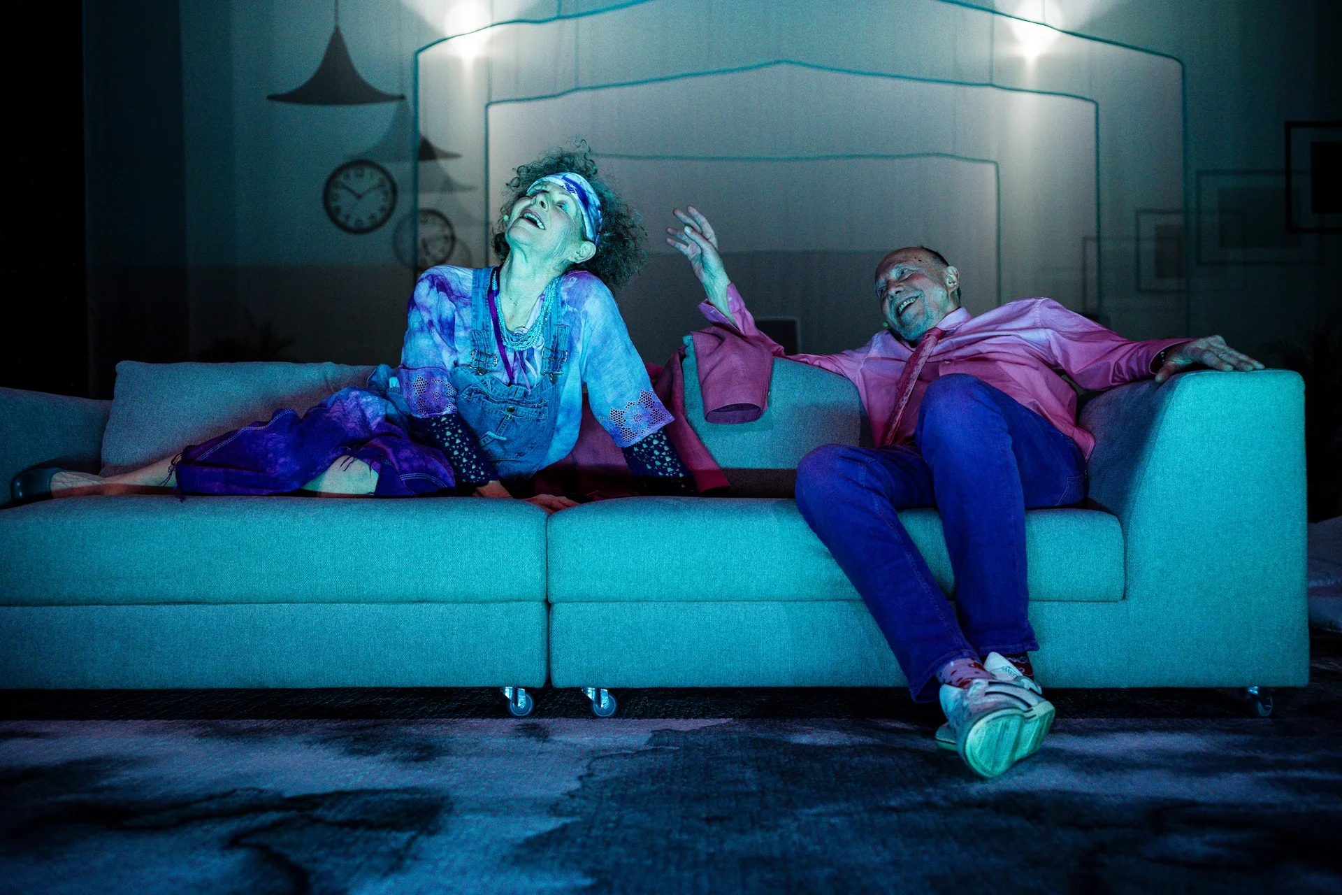 Zdjęcie ze spektaklu Dom niespokojnej starości. Kobieta i mężczyzna siedzą na jasnej kanapie. Ubrani są w kolorowe stroje.