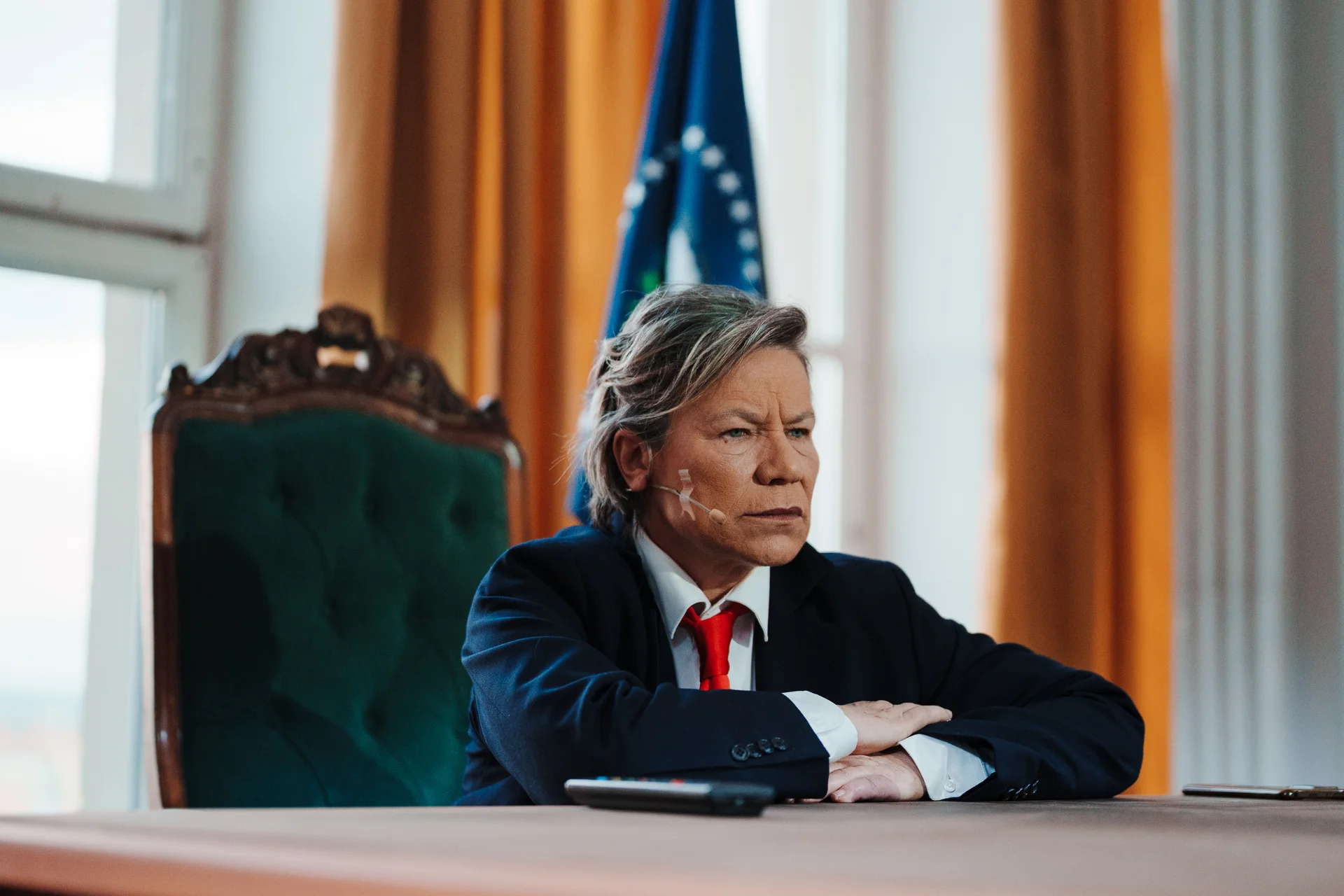 Aktorka w garniturze i czerwonym krawacie siedzi w zielonym eleganckim fotelu oparta przedramionami o blat biurka. W tle żółte zasłony i granatowa flaga