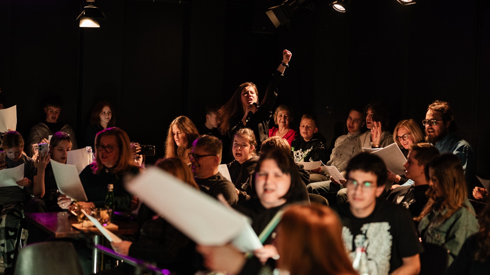 fot. Piotr Nykowski Grupa osób siedząca z kartkami w Teatrze Małym. Część z nich ma w dłoniach kartki z tekstem. Śpiewają. Nad nimi góruje wybijająca się postać kobieca z dłonią uniesioną do góry.