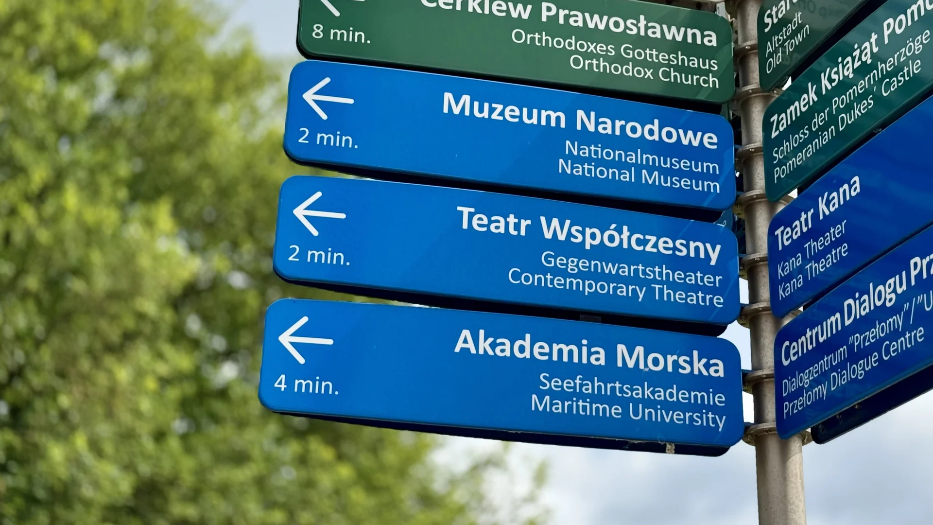zdjęcie drogowskazu. tabliczki są w kolorach niebieskich i zielonych. Wskazują na popularne miejsca w Szczecinie - Teatr Współczesny, Muzeum Narodowe, Akademię Morską itd.